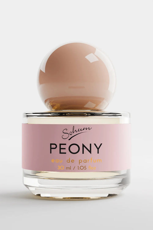 A round glass bottle of Sohum Peony eau de parfum with a blush-toned spherical cap and a soft pink label, featuring black and golden-yellow text for a fresh, feminine look.