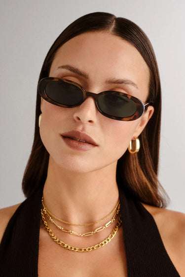Woman wearing sunglasses and gold necklaces against a plain background