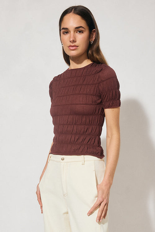 Woman wearing a brown ribbed sweater and white pants against a plain background