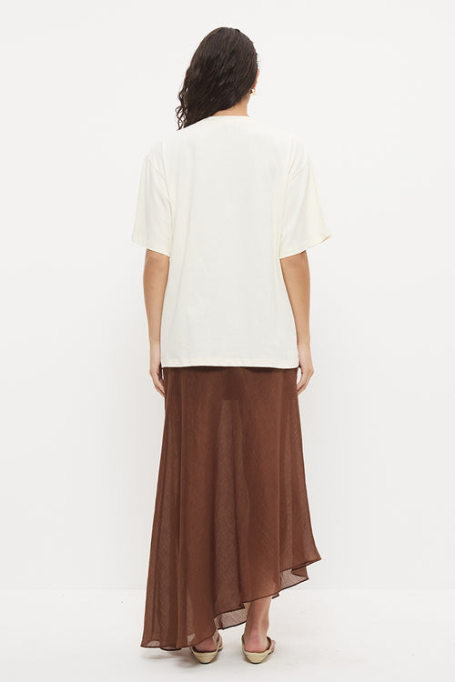 A woman viewed from the back wearing a cream short-sleeve top and a brown high-low midi skirt, standing against a plain white studio background.