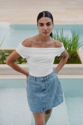 model wears an off the shoulder white top and a blue denim mini skirt by a pool