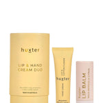A stylishly arranged Huxter lip and hand cream duo set sits on stacked sandstone tiles against a warm-toned background. The set includes a pale yellow tube of hand cream, a white lip balm labelled "Wild Berries & Citrus," and a matching yellow gift box, creating a visually soothing and natural aesthetic.