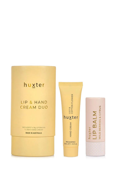 A stylishly arranged Huxter lip and hand cream duo set sits on stacked sandstone tiles against a warm-toned background. The set includes a pale yellow tube of hand cream, a white lip balm labelled 