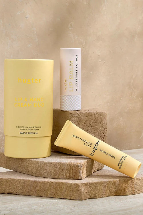 A stylishly arranged Huxter lip and hand cream duo set sits on stacked sandstone tiles against a warm-toned background. The set includes a pale yellow tube of hand cream, a white lip balm labelled 
