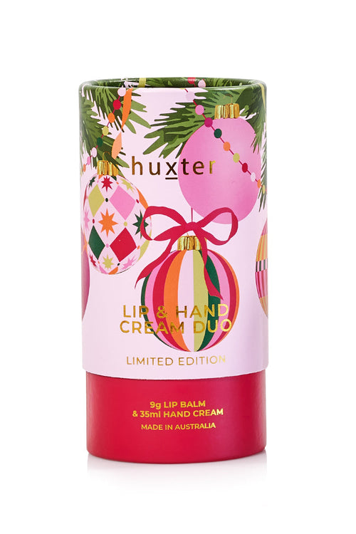 A cylindrical product packaging for a 'Huxter' Lip & Hand Cream Duo in a limited edition holiday theme. The packaging features a light pink upper section decorated with colorful Christmas ornaments, pine branches, and a red ribbon, and a solid red bottom section containing product details.