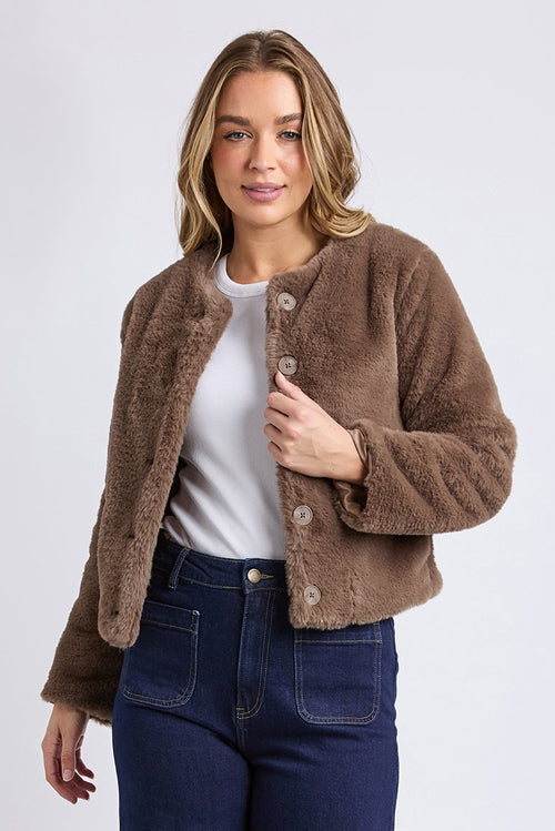 Woman wearing a brown jacket, white shirt, blue jeans, and white sneakers on a light gray background