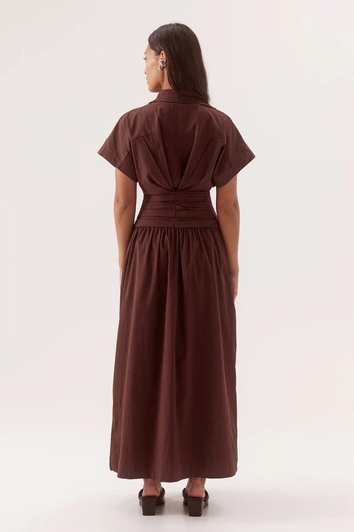 A woman viewed from the back wearing a dark brown short-sleeve midi dress with a gathered waist and pleated back detail, paired with black heels, against a plain light background.
