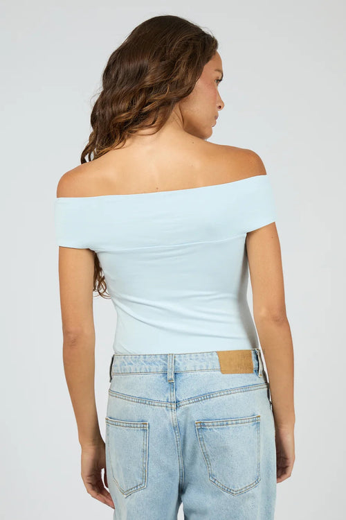 A woman shown from the back wearing a light blue off-the-shoulder fitted top and light-wash blue jeans against a plain light gray studio background.