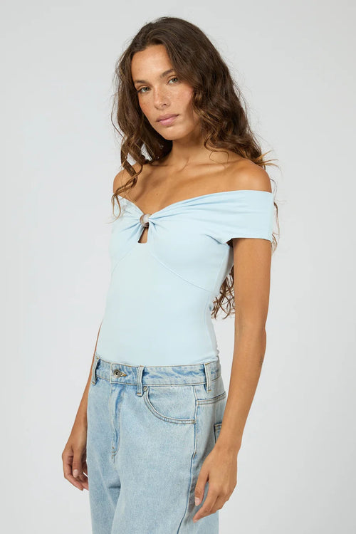 A woman modeling a light blue off-the-shoulder knit top with a front knot detail, paired with light-wash jeans, against a plain light gray studio background.