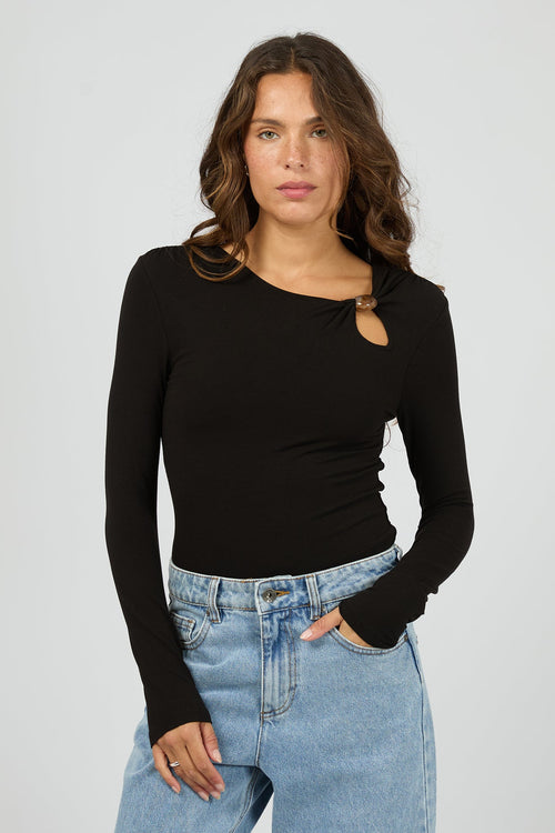 Woman wearing a black long-sleeve top with a cutout design and light blue jeans on a gray background