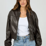 Woman wearing a brown leather jacket, white shirt, and blue jeans on a gray background