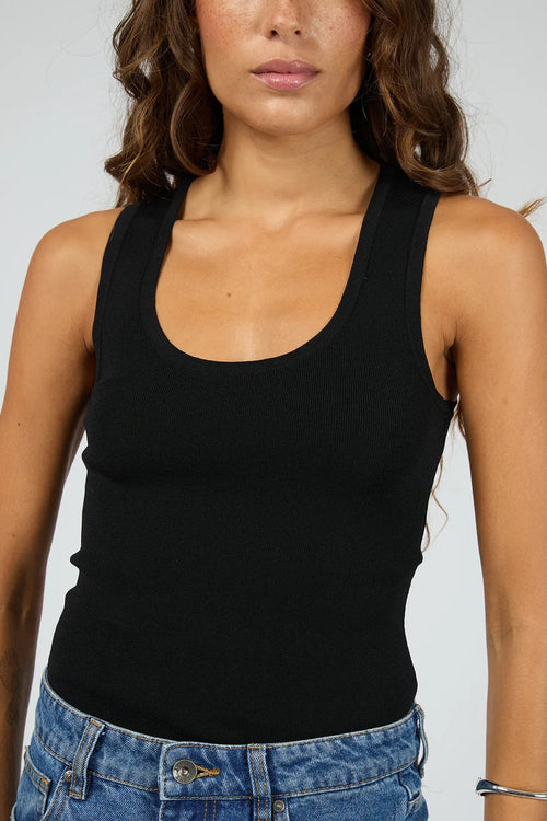 A woman wearing a black scoop-neck tank top and blue jeans, cropped from the nose down, against a light gray studio background.
