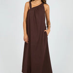 A woman modeling a dark brown one-shoulder maxi dress with a side pocket, paired with brown sandals, against a light gray studio background.