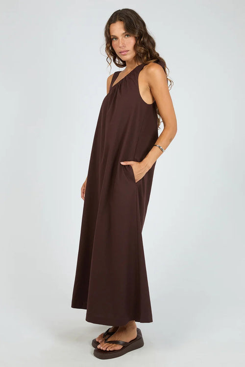 A woman modeling a sleeveless, dark brown maxi dress with a draped neckline and side pockets, standing against a light gray studio background.