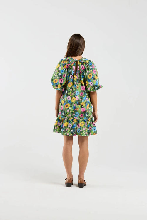 A woman viewed from behind wearing a short puff-sleeve dress with a colorful floral print on a green background, paired with dark brown shoes, against a plain white studio backdrop.