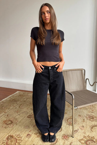 A woman stands indoors wearing a fitted dark charcoal short-sleeve crop top and very wide-leg black jeans, paired with black flats. She is posed in front of a white wall and a woven chair, with a patterned rug on the floor.