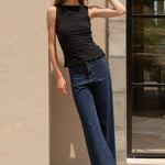Woman in black top and blue jeans standing against a beige wall with a glass door in the background.