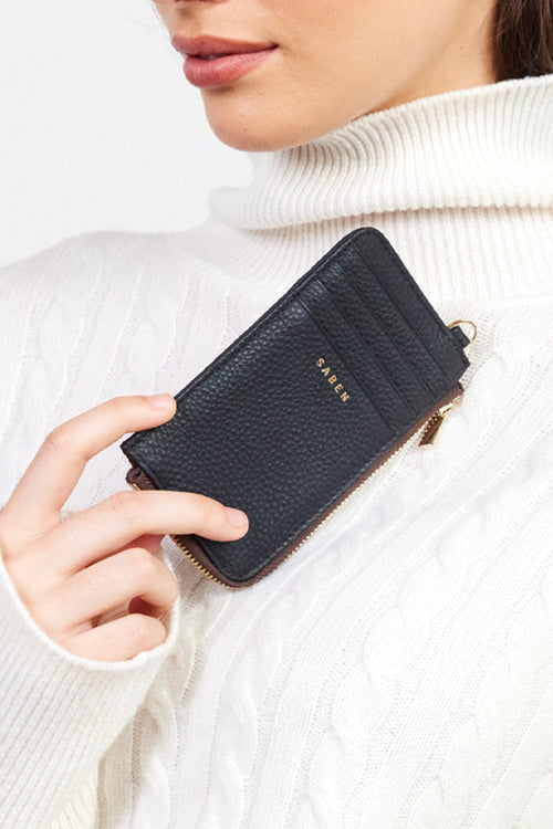 Person holding a black wallet with a brand logo, wearing a white sweater.