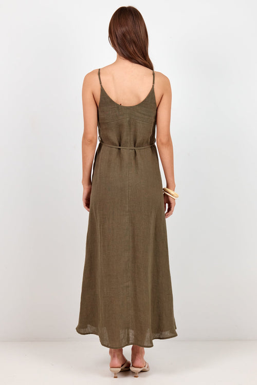 A woman viewed from the back wearing a sleeveless olive green maxi dress with thin straps and a tie at the waist, standing against a plain light gray studio background.