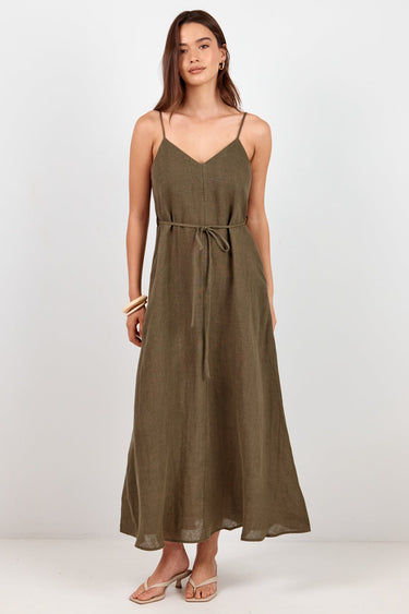 A woman modeling a long olive green spaghetti-strap maxi dress with a tie waist, standing against a plain white studio background.