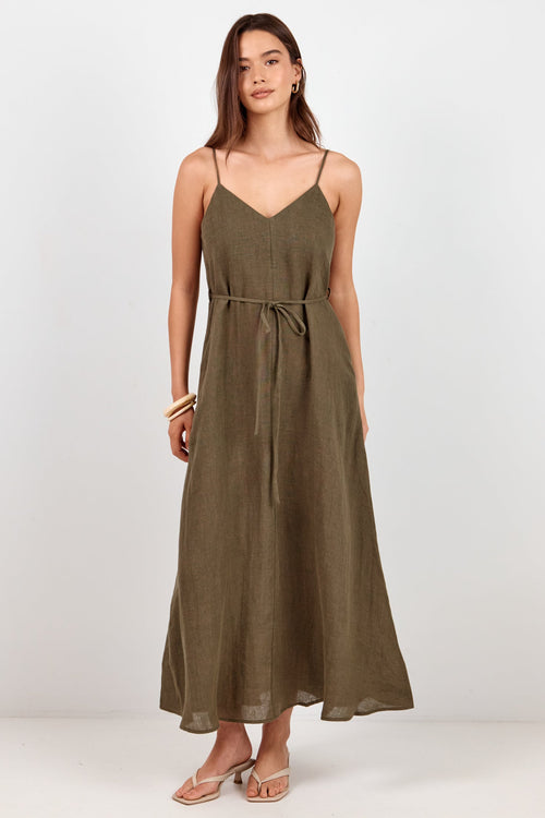 A woman modeling a long olive green spaghetti-strap maxi dress with a tie waist, standing against a plain white studio background.
