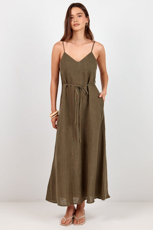A woman modeling a sleeveless olive green maxi dress with thin spaghetti straps, a V-neckline, side pockets, and a matching fabric tie belt at the waist, set against a plain white studio background.