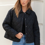 Woman wearing a black jacket and blue jeans standing against a white building.