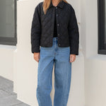 Woman wearing a black quilted jacket and blue jeans standing in front of a building.