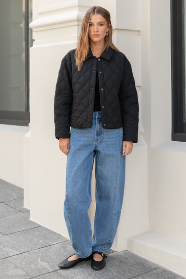 Woman wearing a black quilted jacket and blue jeans standing in front of a building.