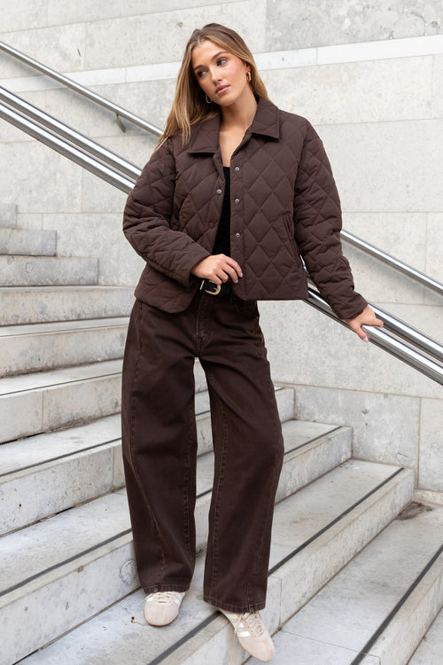 Woman in a brown outfit standing on a staircase with a light gray wall background