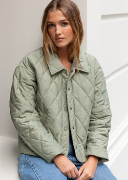 Woman wearing a green quilted jacket and blue jeans against a white wall.