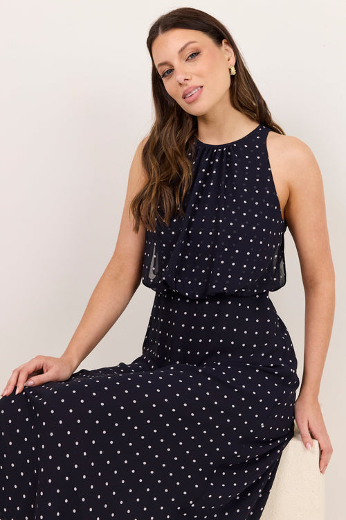 A woman wearing a sleeveless navy maxi dress with a white polka dot pattern, sitting on a white stool against a plain off-white background.