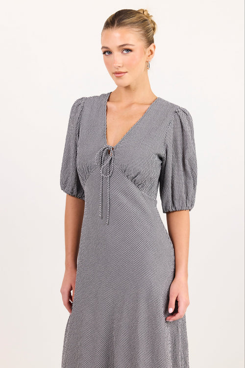 A woman modeling a gray and white micro-checkered midi dress with puff sleeves, a V-neckline, and a front tie detail, set against a plain off-white studio background.