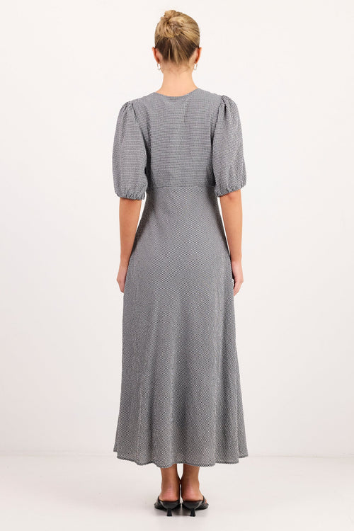 A woman viewed from the back wearing a gray midi dress with short puff sleeves and black flat shoes, standing against a plain white studio background.