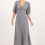 A woman modeling a long gray midi dress with puff sleeves, a V-neckline, and a tie detail at the bust, photographed against a plain light studio background.