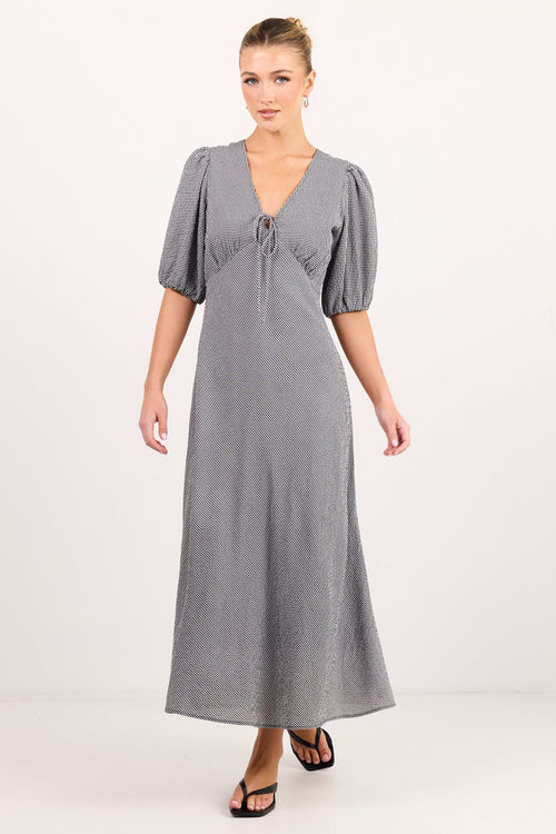 A woman modeling a long gray midi dress with puff sleeves, a V-neckline, and a tie detail at the bust, photographed against a plain light studio background.