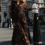 Woman in a long dress standing next to a street sign in an urban setting with classical architecture.