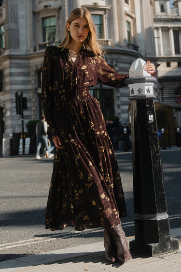 Woman in a long dress standing next to a street sign in an urban setting with classical architecture.
