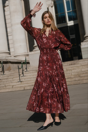 Woman in a red floral dress waving in front of a classical building.