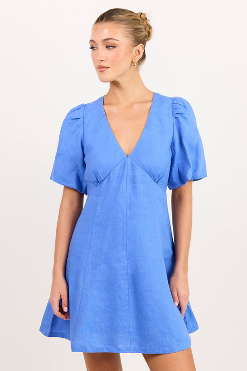 A woman modeling a short-sleeve blue dress with a deep V-neckline, puffed sleeves, and a flared skirt against a plain white studio background.