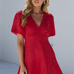 A woman wearing a short-sleeve red mini dress and black flip-flops stands on a white tiled patio with white walls and dark green trees in the background.