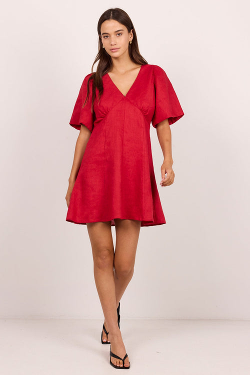 A woman modeling a short red wrap-style dress with short puff sleeves and a V-neckline, standing against a plain light studio background.