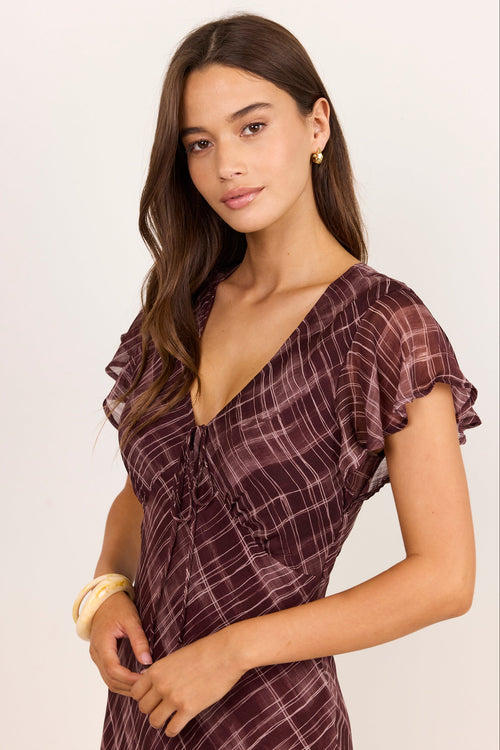 A woman modeling a short-sleeve wrap dress with a ruffled V-neckline and a subtle plaid pattern in dark plum and brown tones, set against a plain light background.