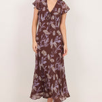 A woman modeling a brown midi dress with a purple floral pattern, ruffled cap sleeves, and a V-neckline, standing against a plain off-white studio background.