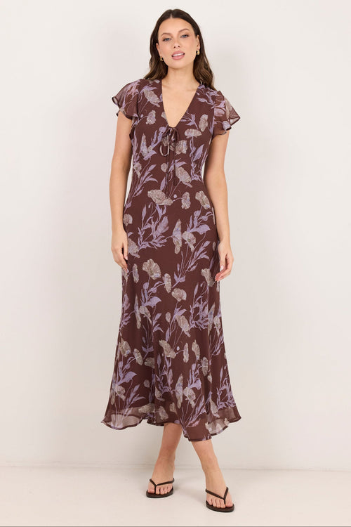 A woman modeling a brown midi dress with a purple floral pattern, ruffled cap sleeves, and a V-neckline, standing against a plain off-white studio background.