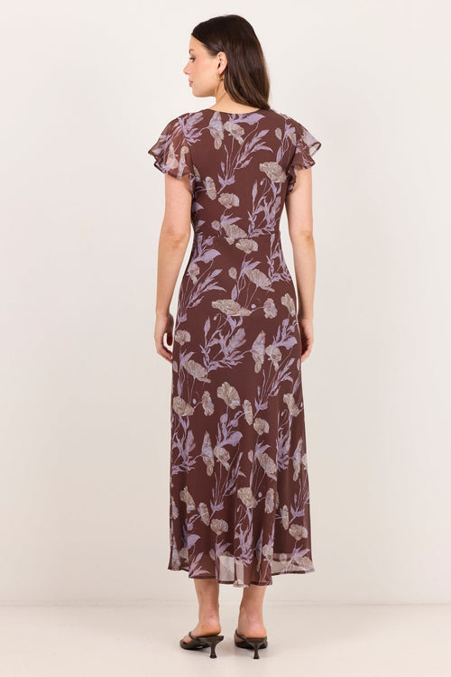 A woman stands with her back to the camera wearing a brown midi dress with a light purple floral pattern, ruffled cap sleeves, and black pointed-toe heels against a plain off-white studio background.