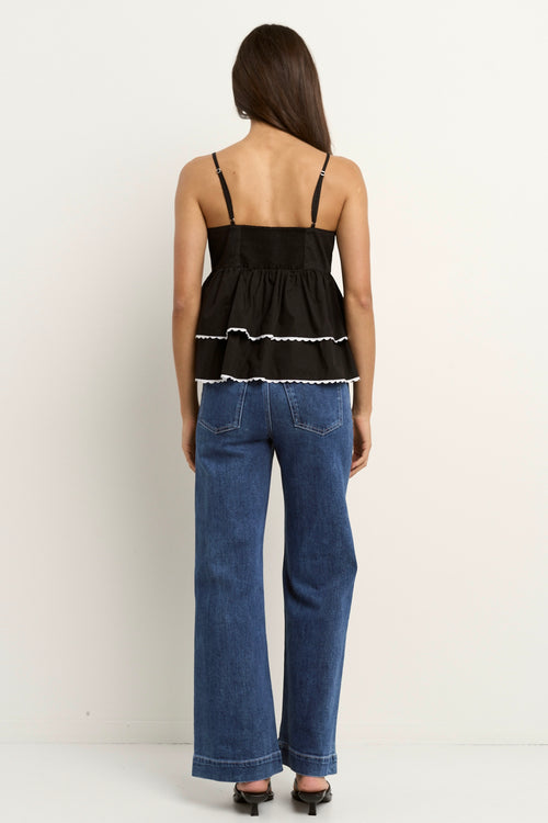 Woman wearing a black tiered ruffle cami top with white scalloped hem detail and blue jeans.