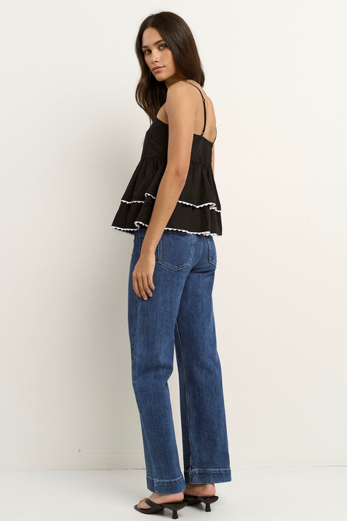 Woman wearing a black tiered ruffle cami top with white scalloped hem detail and blue jeans.