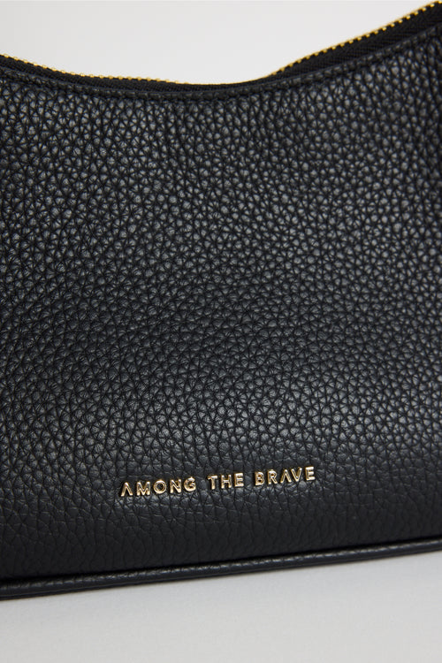 Close-up of a black pebbled leather pouch with gold zipper trim and gold 'AMONG THE BRAVE' branding on a light gray background.