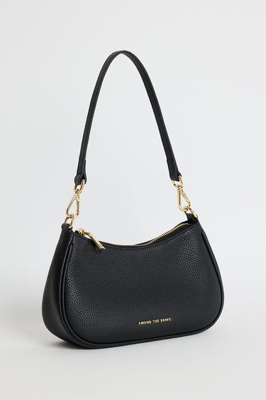 Black pebble-textured leather shoulder bag with gold hardware, featuring a curved silhouette, double adjustable straps and top zip closure. Stylish small black handbag from Among the Brave, ideal for everyday wear.